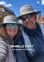 A man and a woman taking a selfie outdoors under a blue sky, both wearing gray wide-brim shield hats with mesh neck covers and sunglasses. White text overlay at the bottom reads 'SHIELD HAT BY SORIA TACTICAL'.
